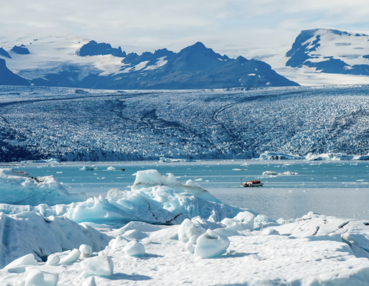 A photo of a small boat in a waterway surrounded by iceburgs and mountains.